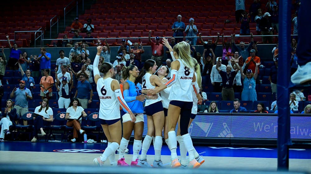 Auburn volleyball players celebrate on the court after a point, huddled together in excitement, while fans in the stands cheer and clap in the background.
