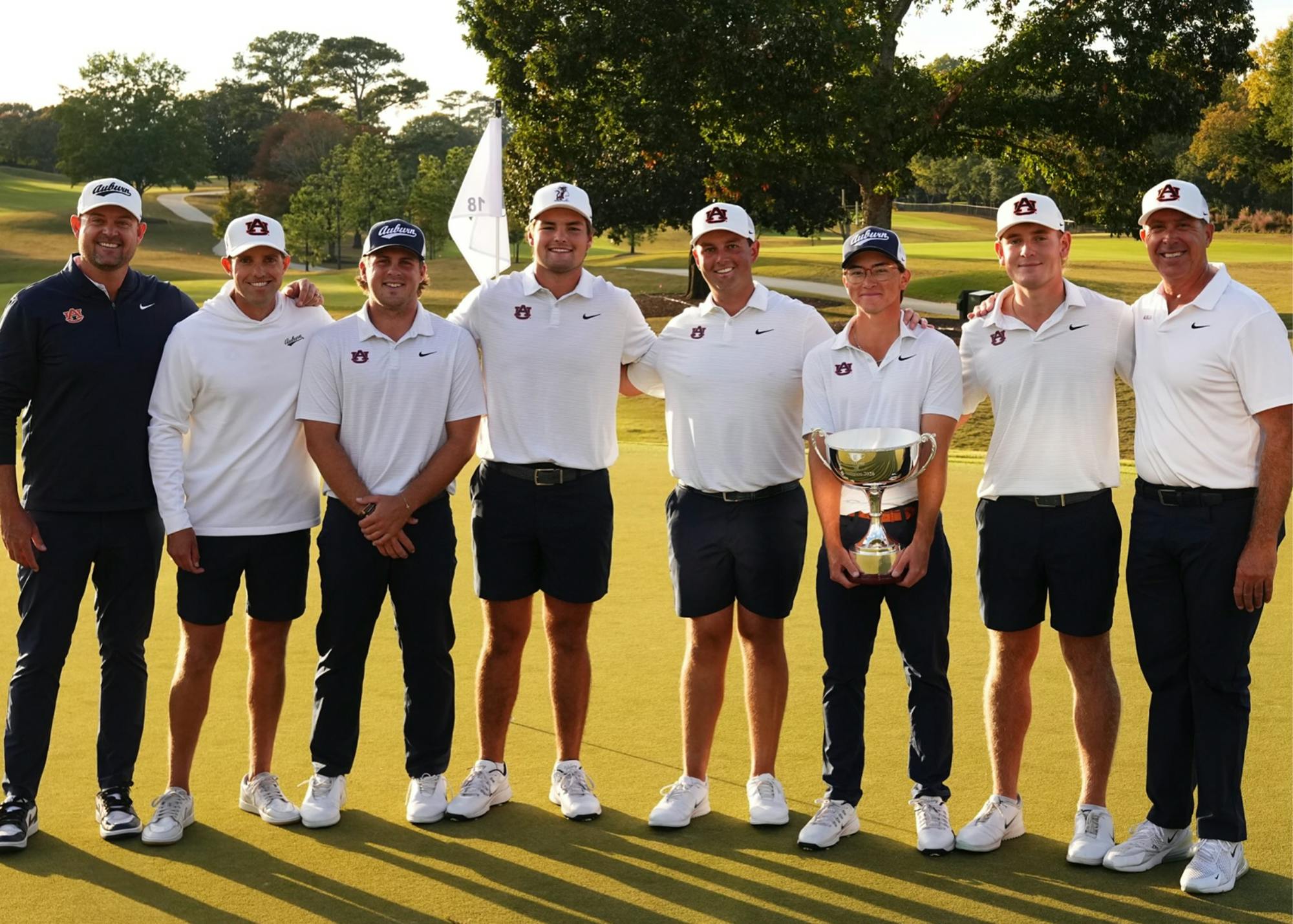 Auburn men’s golf players and coaches pose together on the golf course following their East Lake Cup victory, with one player holding the championship trophy. The group wears Auburn-branded white and navy golf apparel and hats, standing on the green with the course and trees in the background.