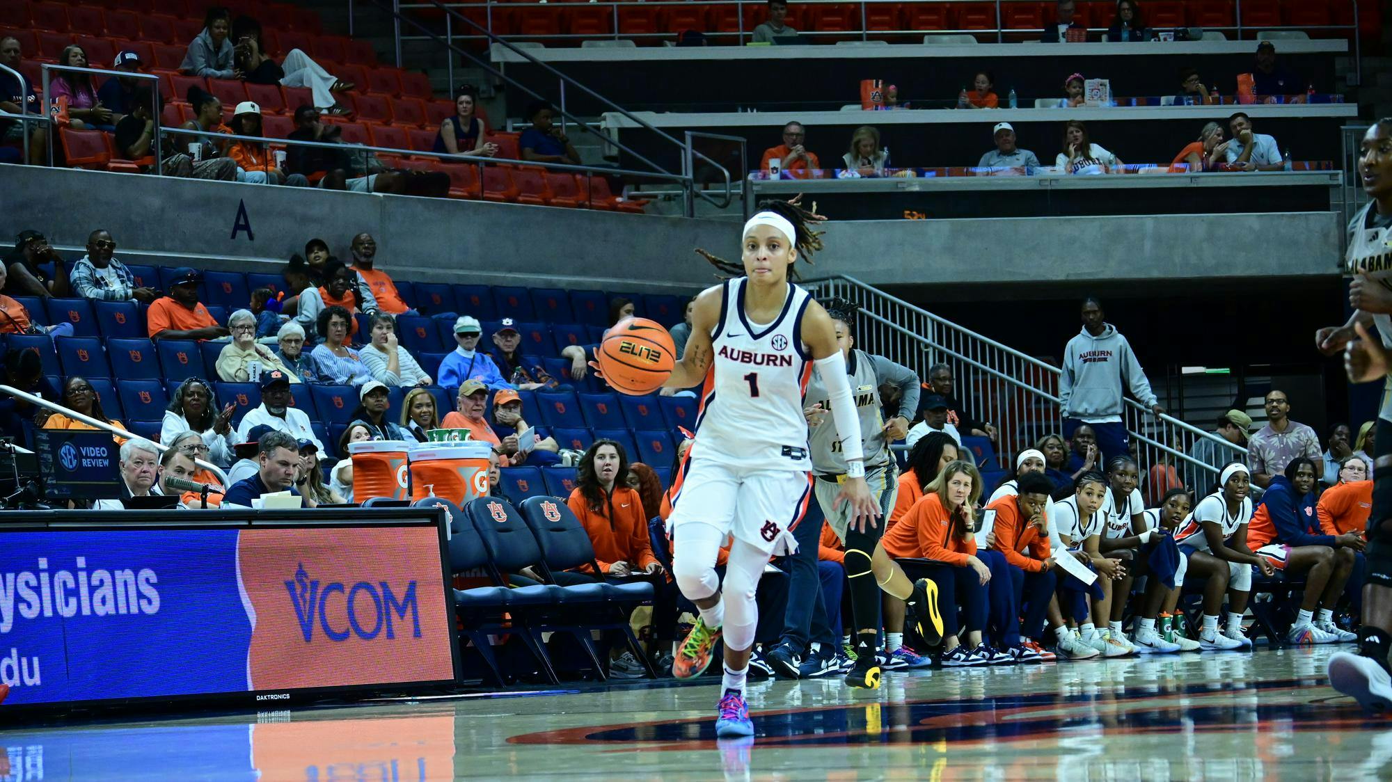 An Auburn women’s basketball player wearing a white uniform with number 1 dribbles the ball up the court during a game. The Auburn bench and coaching staff watch intently from the sideline, while fans in orange and blue fill the stands behind them. The court and arena lighting highlight the action as she advances the play.