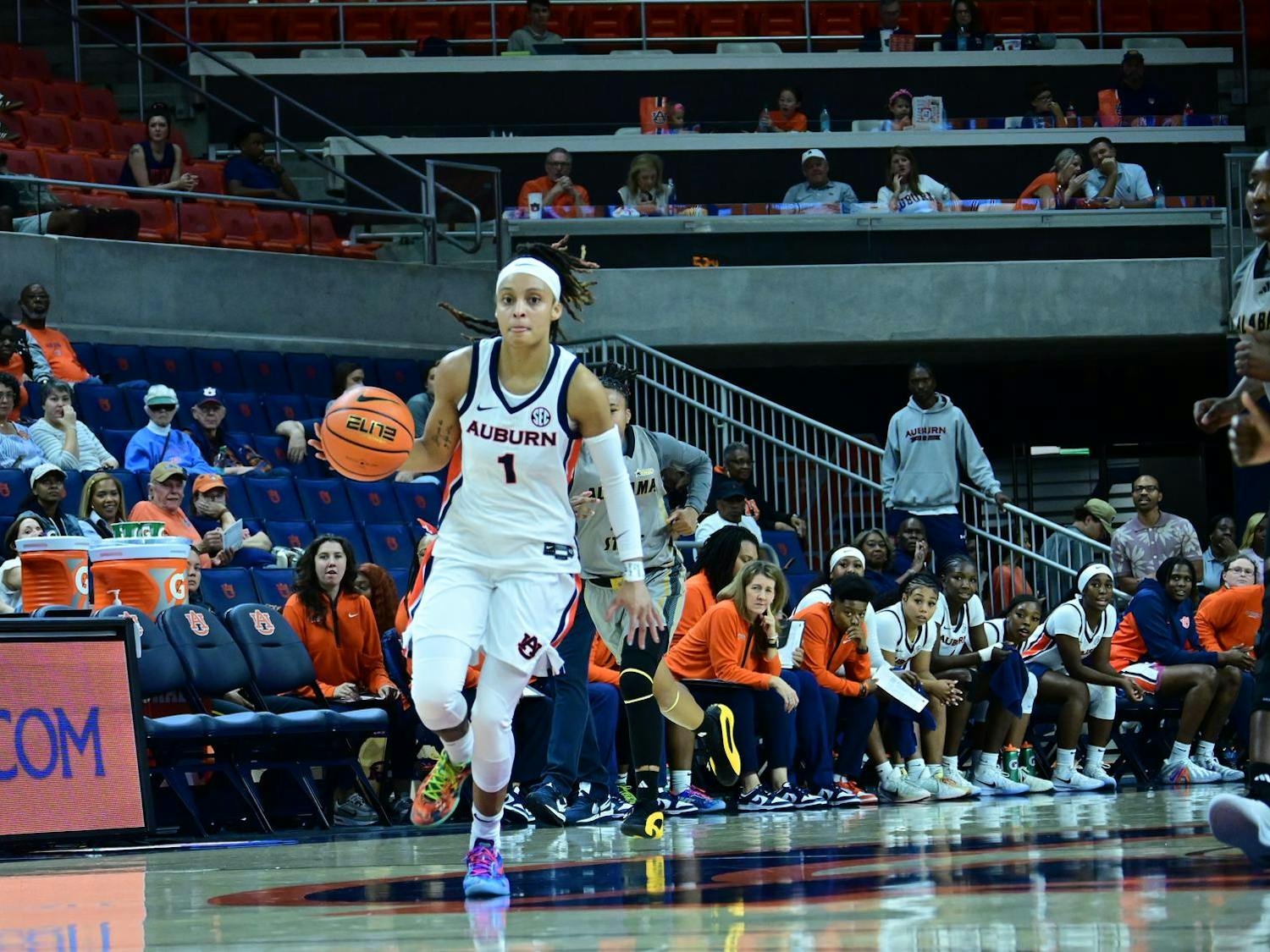 An Auburn women’s basketball player wearing a white uniform with number 1 dribbles the ball up the court during a game. The Auburn bench and coaching staff watch intently from the sideline, while fans in orange and blue fill the stands behind them. The court and arena lighting highlight the action as she advances the play.