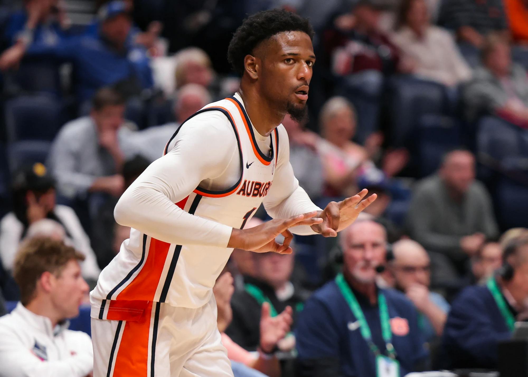 An Auburn men’s basketball player in a white jersey with orange and navy trim gestures with both hands during a game, appearing to signal a play or celebrate, while a blurred crowd and sideline staff watch from the background.
