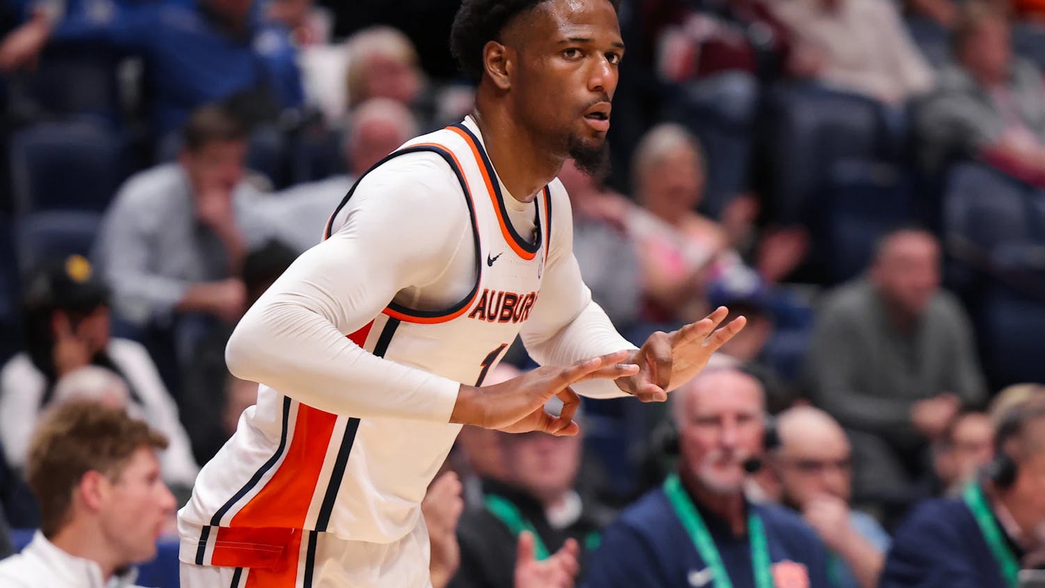 An Auburn men’s basketball player in a white jersey with orange and navy trim gestures with both hands during a game, appearing to signal a play or celebrate, while a blurred crowd and sideline staff watch from the background.