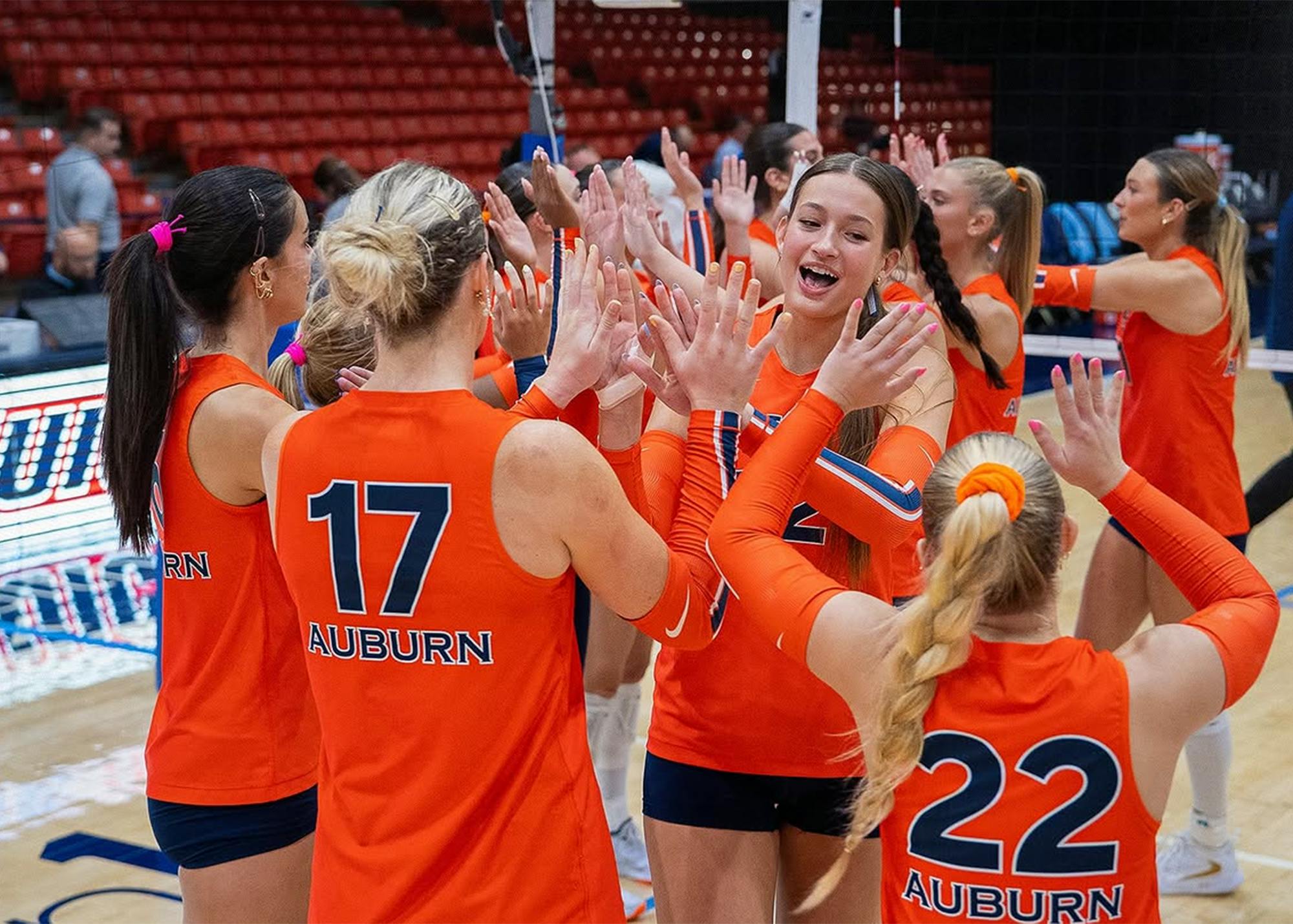 Auburn volleyball players in orange jerseys celebrate together with high fives during a match, with players wearing Nos. 17 and 22 in the foreground and the court and stands in the background.
