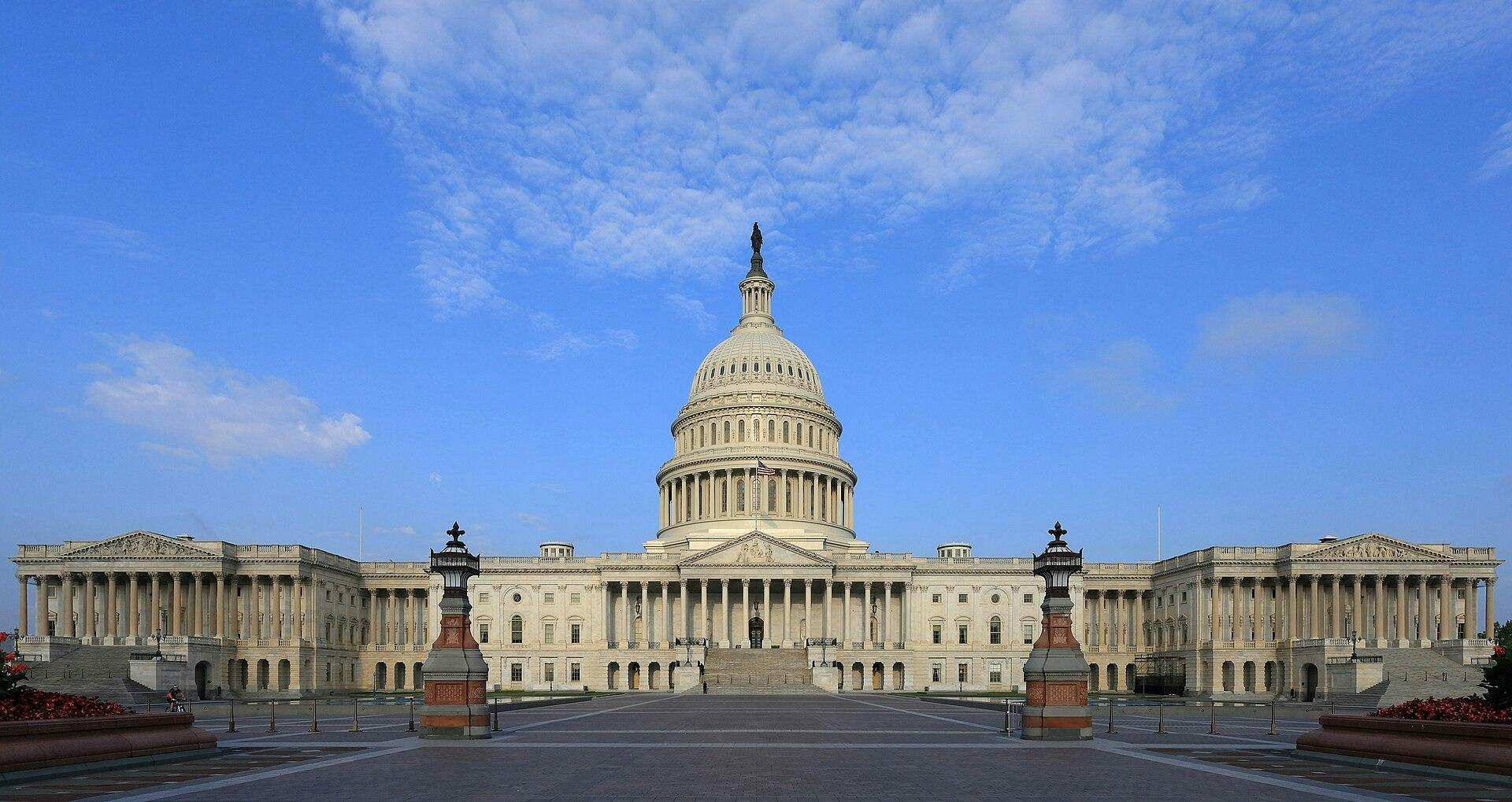 The east side of the U.S. Capitol building fills the center of the photo, with its dome rising above a broad plaza under a bright blue, cloud-filled sky.