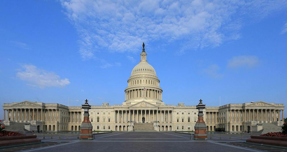 The east side of the U.S. Capitol building fills the center of the photo, with its dome rising above a broad plaza under a bright blue, cloud-filled sky.