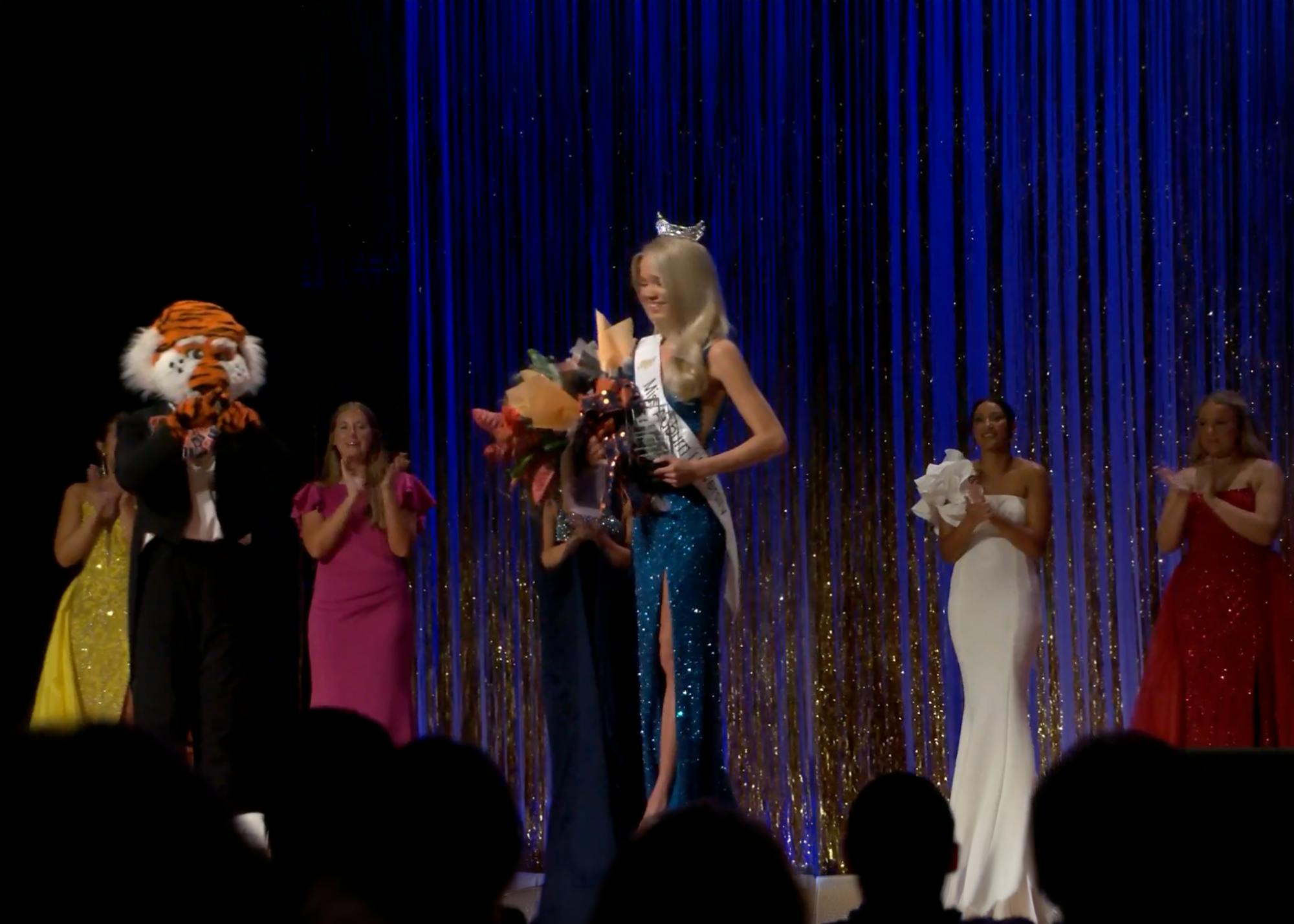 Emma Bishop wears a crown, sash and blue sequined gown while holding flowers after winning Miss Auburn University on Oct. 2. Fellow contestants and Auburn mascot Aubie applaud onstage