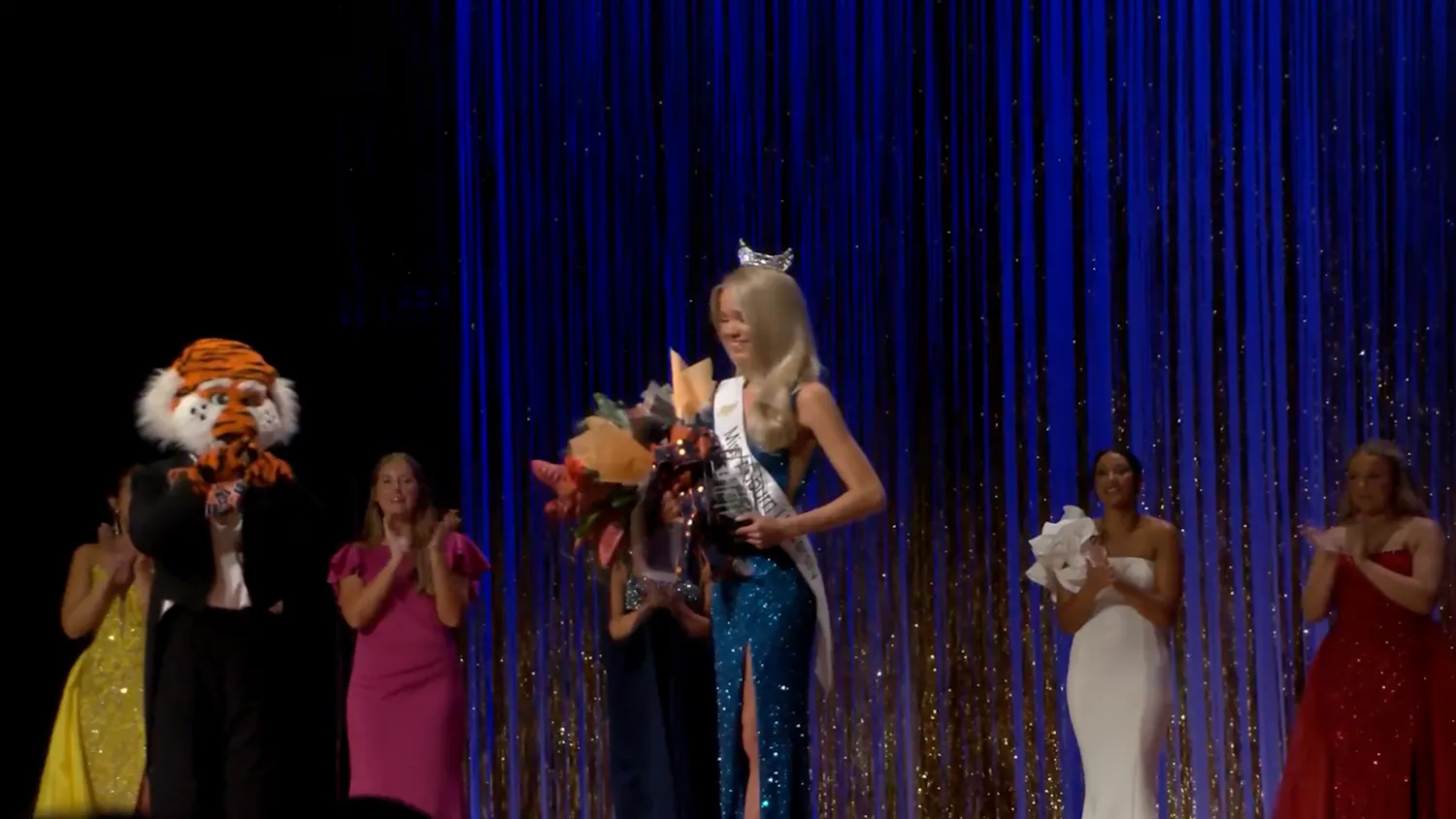 Emma Bishop wears a crown, sash and blue sequined gown while holding flowers after winning Miss Auburn University on Oct. 2. Fellow contestants and Auburn mascot Aubie applaud onstage