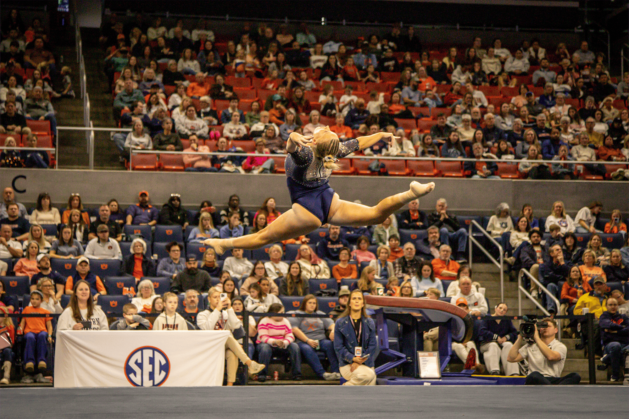 Auburn gymnast in a navy leotard performs a high split leap during a floor routine, arms extended and legs stretched wide in midair, with judges and a large crowd watching inside an arena.