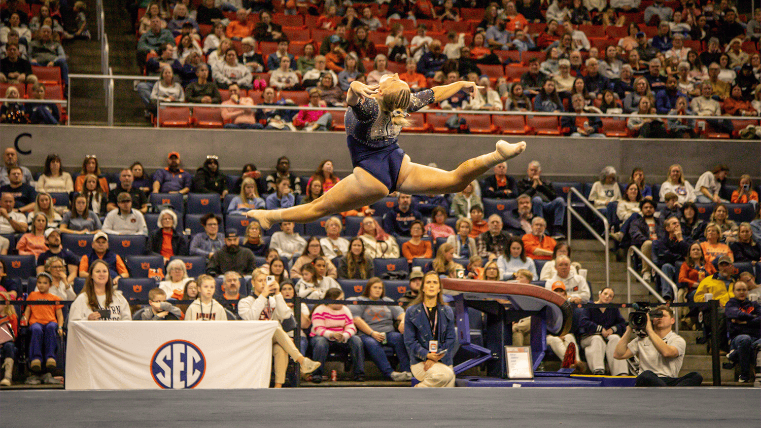 Auburn gymnast in a navy leotard performs a high split leap during a floor routine, arms extended and legs stretched wide in midair, with judges and a large crowd watching inside an arena.