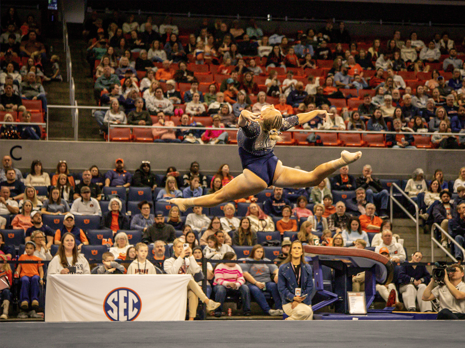 Auburn gymnast in a navy leotard performs a high split leap during a floor routine, arms extended and legs stretched wide in midair, with judges and a large crowd watching inside an arena.