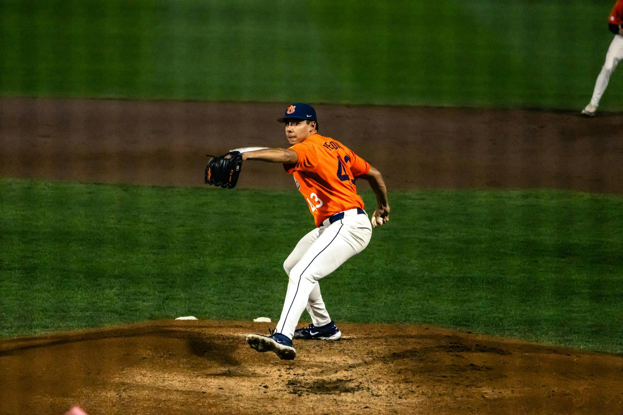 An Auburn baseball pitcher wearing an orange jersey and gray pants delivers a pitch from the mound during a night game. His glove is extended forward and his body is mid-rotation, with the dirt mound beneath him and the green outfield grass in the background.