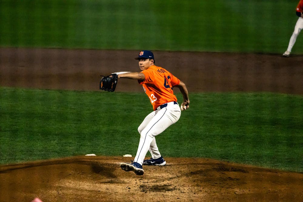 An Auburn baseball pitcher wearing an orange jersey and gray pants delivers a pitch from the mound during a night game. His glove is extended forward and his body is mid-rotation, with the dirt mound beneath him and the green outfield grass in the background.