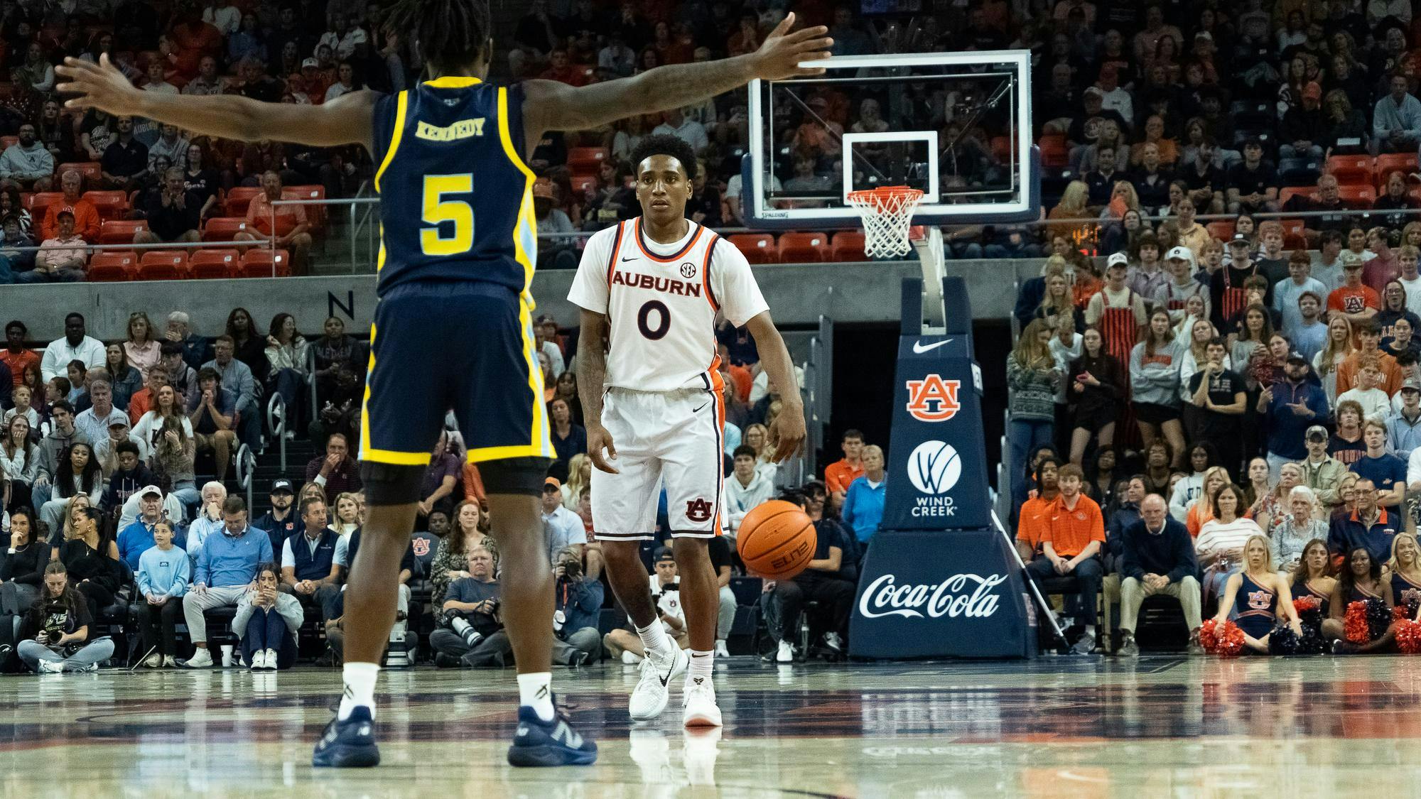 An Auburn basketball player wearing a white jersey with number 0 dribbles the ball near midcourt while facing a defender in a navy and yellow uniform with number 5. The game takes place in a packed arena, with fans, cheerleaders, and photographers watching closely from the sidelines and stands.