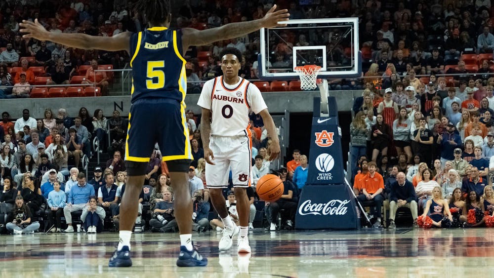 An Auburn basketball player wearing a white jersey with number 0 dribbles the ball near midcourt while facing a defender in a navy and yellow uniform with number 5. The game takes place in a packed arena, with fans, cheerleaders, and photographers watching closely from the sidelines and stands.