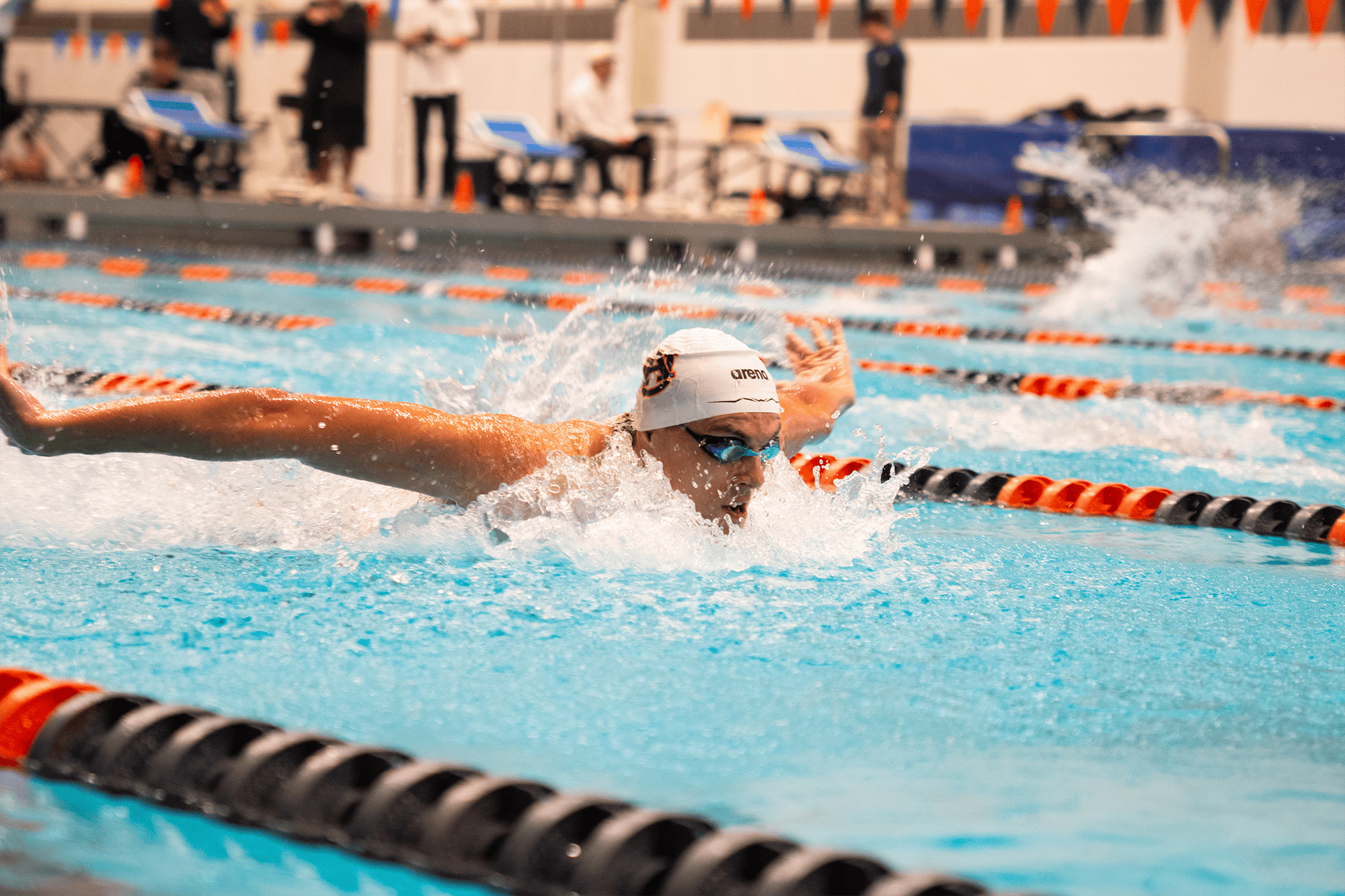 Auburn swimmer wearing a white team cap and goggles performs the butterfly stroke in a lane of an indoor pool, arms extended and water splashing, with lane lines, officials, and spectators blurred in the background.