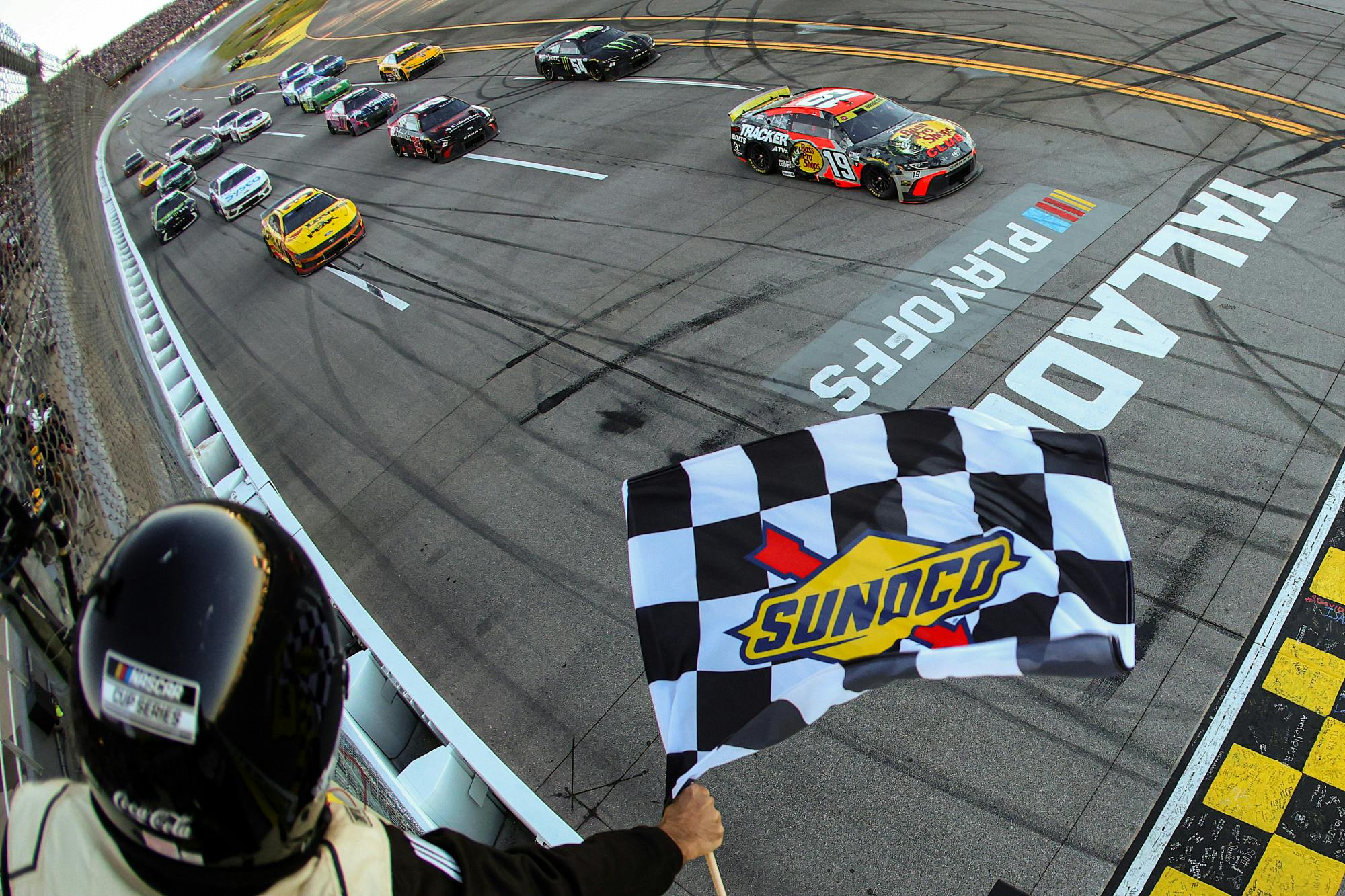 A NASCAR official waves a Sunoco-branded checkered flag as a race car crosses the finish line at Talladega Superspeedway, marking the end of a playoff race, with several other cars following close behind.