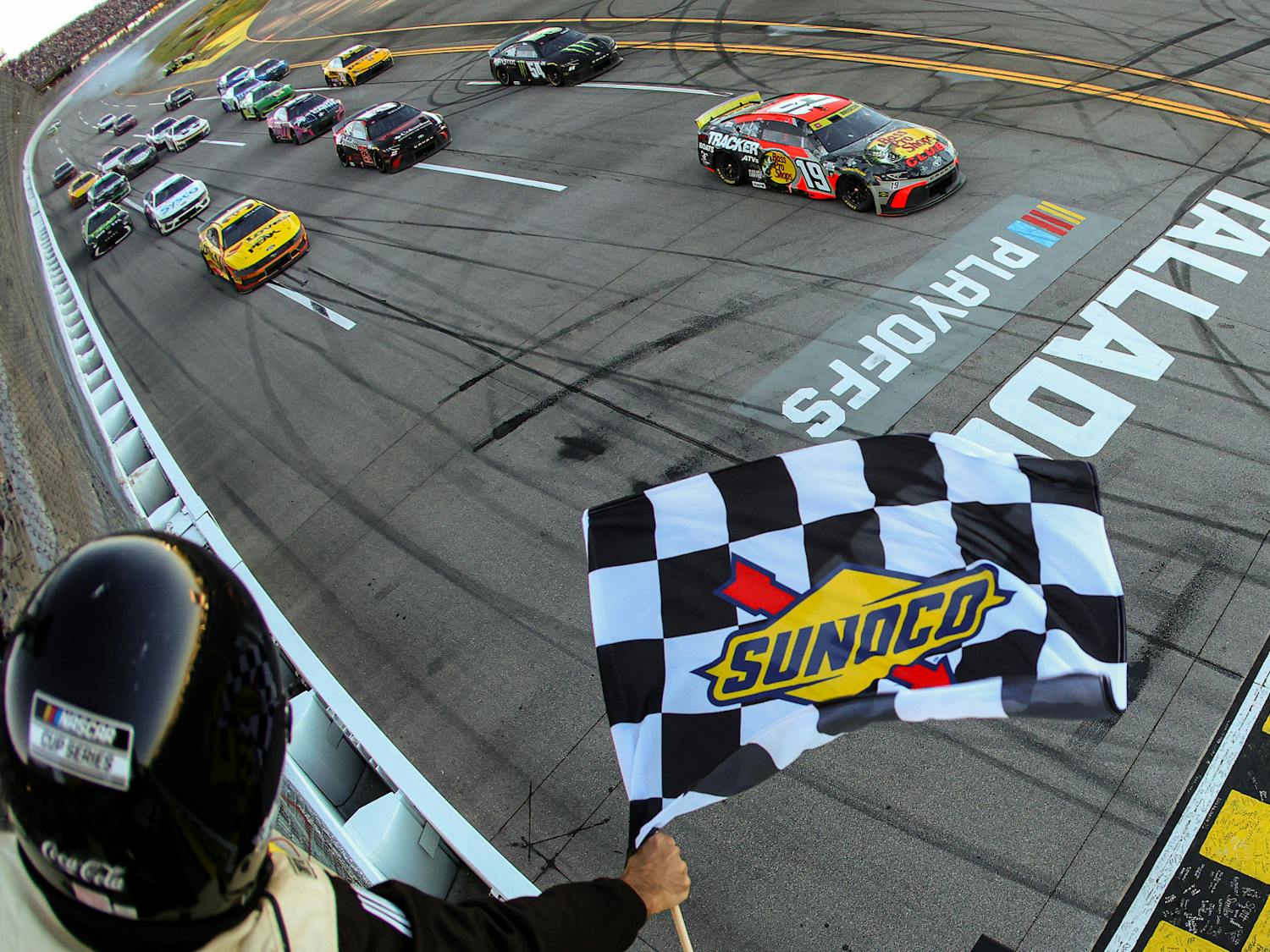 A NASCAR official waves a Sunoco-branded checkered flag as a race car crosses the finish line at Talladega Superspeedway, marking the end of a playoff race, with several other cars following close behind.