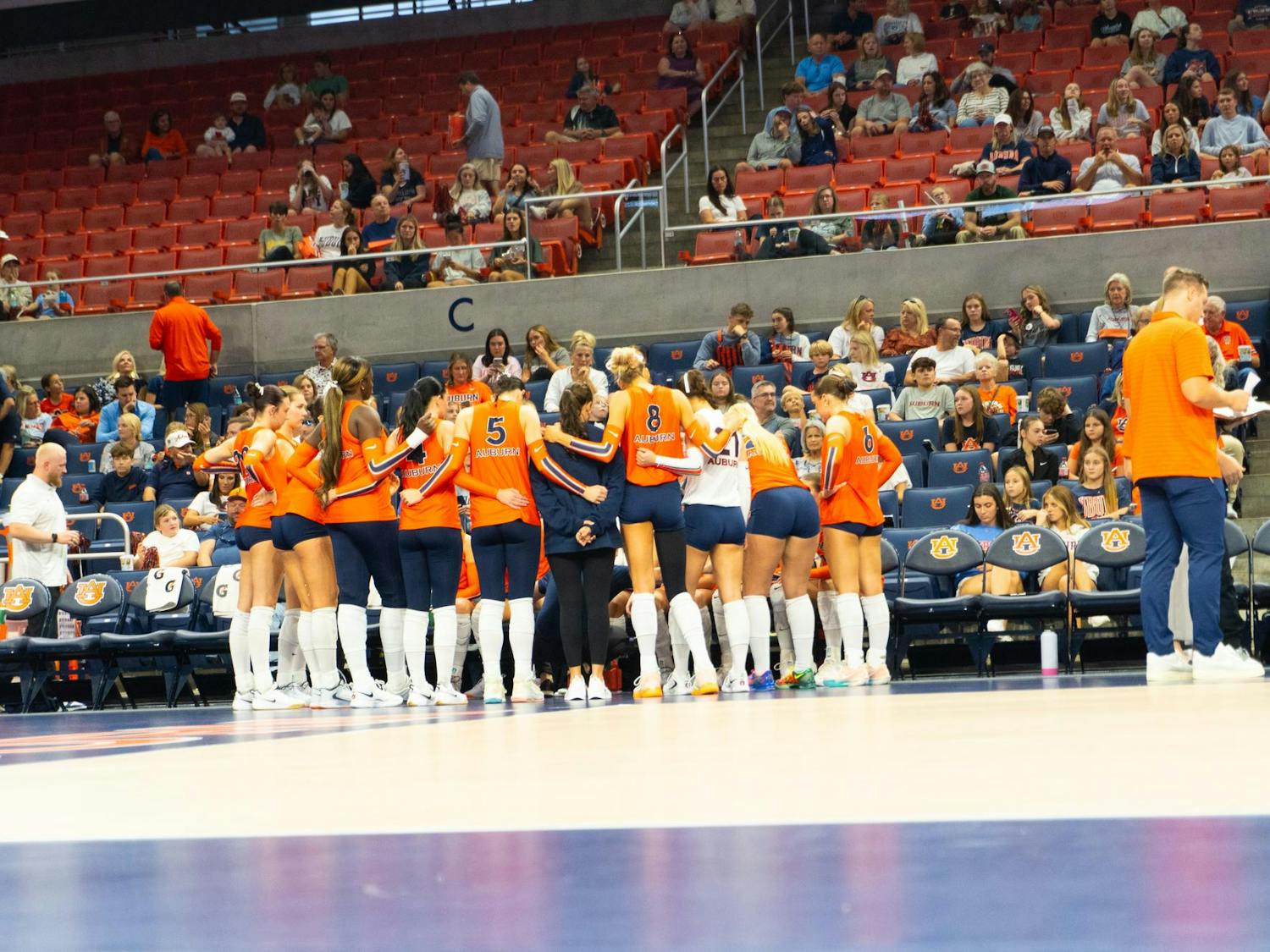 Auburn volleyball players huddle together on the court during a timeout, with coaches standing nearby and fans watching from the stands in Neville Arena.