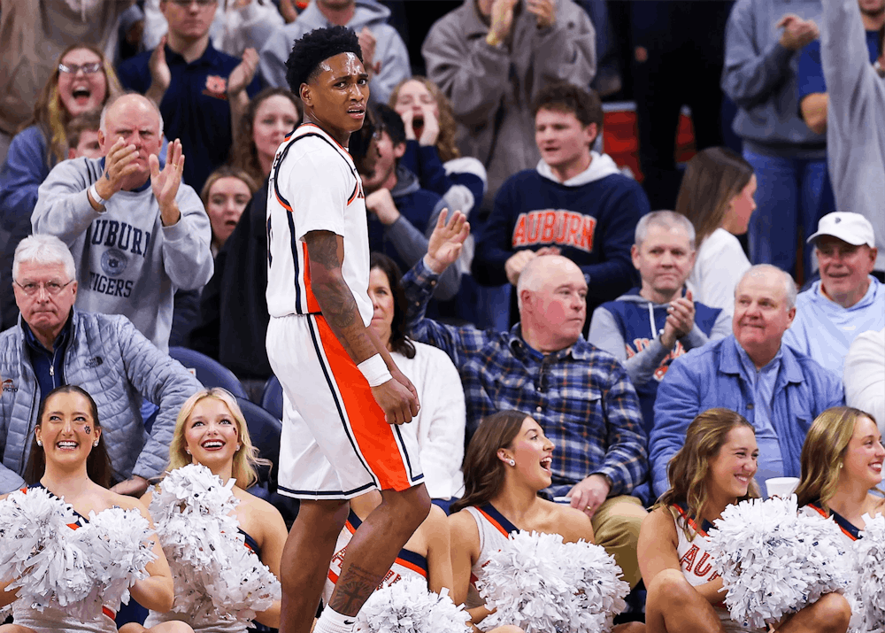 A basketball player in a white and orange Auburn uniform stands near the baseline, looking toward the court with a focused expression as fans behind him cheer and clap. Several Auburn cheerleaders seated along the sideline smile and hold white pom-poms, while the crowd in the background, many wearing Auburn apparel, reacts energetically to the play.