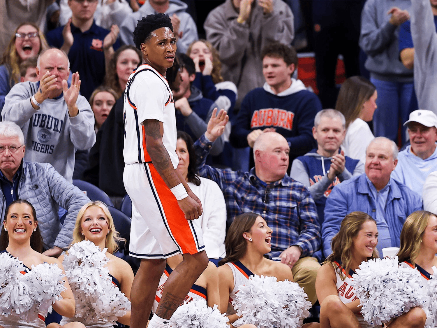 A basketball player in a white and orange Auburn uniform stands near the baseline, looking toward the court with a focused expression as fans behind him cheer and clap. Several Auburn cheerleaders seated along the sideline smile and hold white pom-poms, while the crowd in the background, many wearing Auburn apparel, reacts energetically to the play.