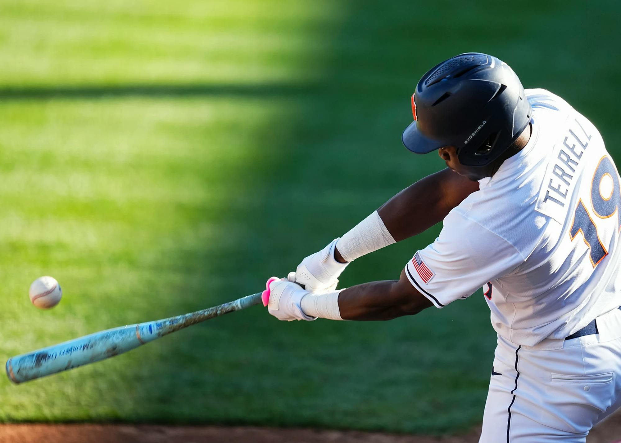A baseball player in a white uniform with “TERRELL” and the number 19 on the back swings a bat, making contact with a pitch as the ball flies off to the left. He wears a dark helmet and batting gloves, and the action is captured mid-swing against a blurred green outfield background.
