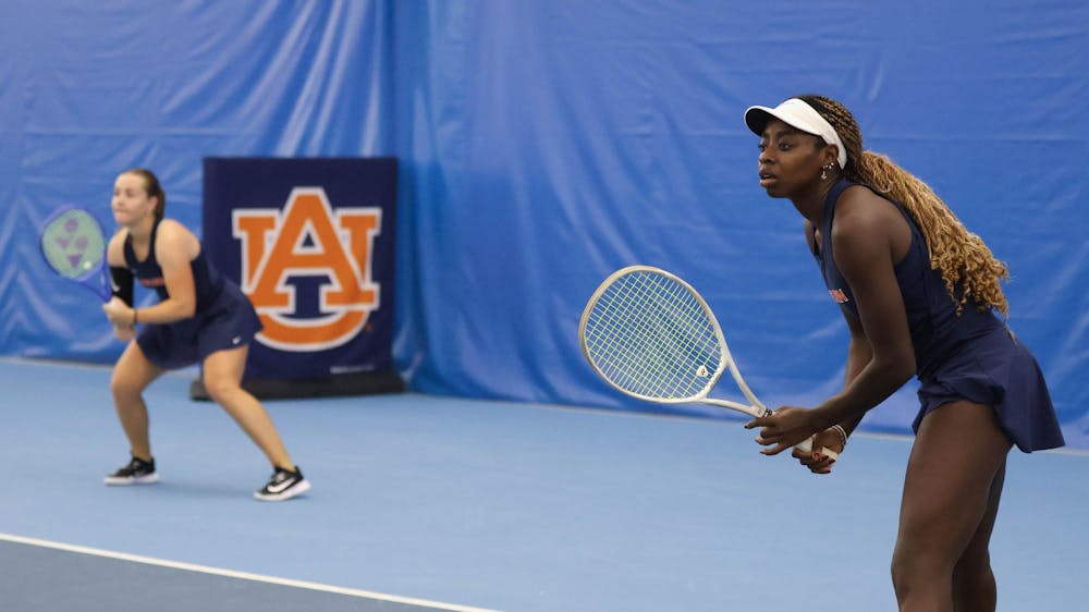 Two women’s tennis players in navy Auburn uniforms prepare to receive a serve on an indoor blue court. The player in the foreground, wearing a white visor, holds her racket ready, while her doubles partner crouches behind her. An Auburn “AU” logo banner is visible in the background.