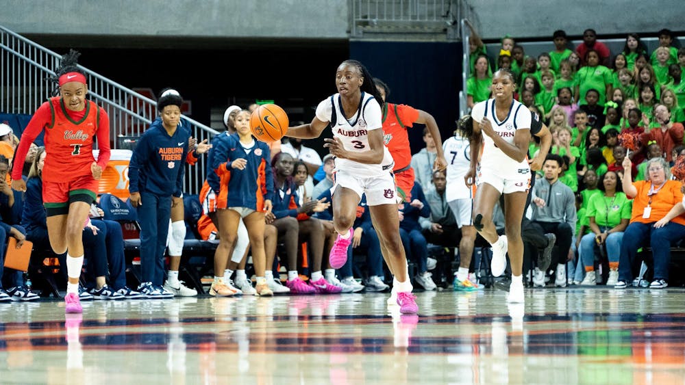 A women’s college basketball player from Auburn, wearing a white uniform, dribbles the ball quickly up the court during a game, with a teammate running alongside her. An opposing player in a red and green uniform trails close behind. The Auburn bench and a crowd of spectators, including a large group wearing bright green shirts, are visible in the background.