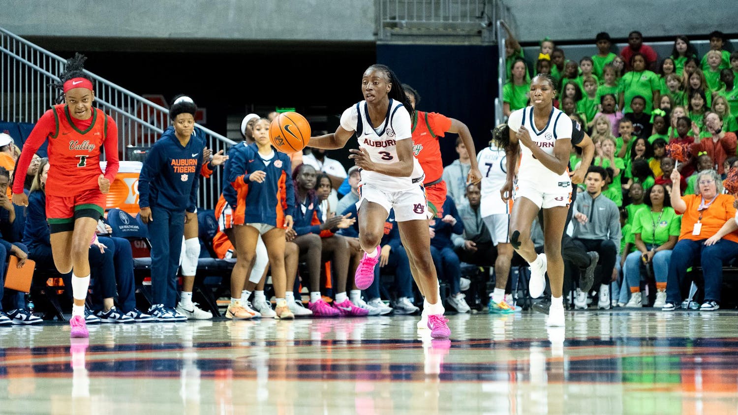 A women’s college basketball player from Auburn, wearing a white uniform, dribbles the ball quickly up the court during a game, with a teammate running alongside her. An opposing player in a red and green uniform trails close behind. The Auburn bench and a crowd of spectators, including a large group wearing bright green shirts, are visible in the background.