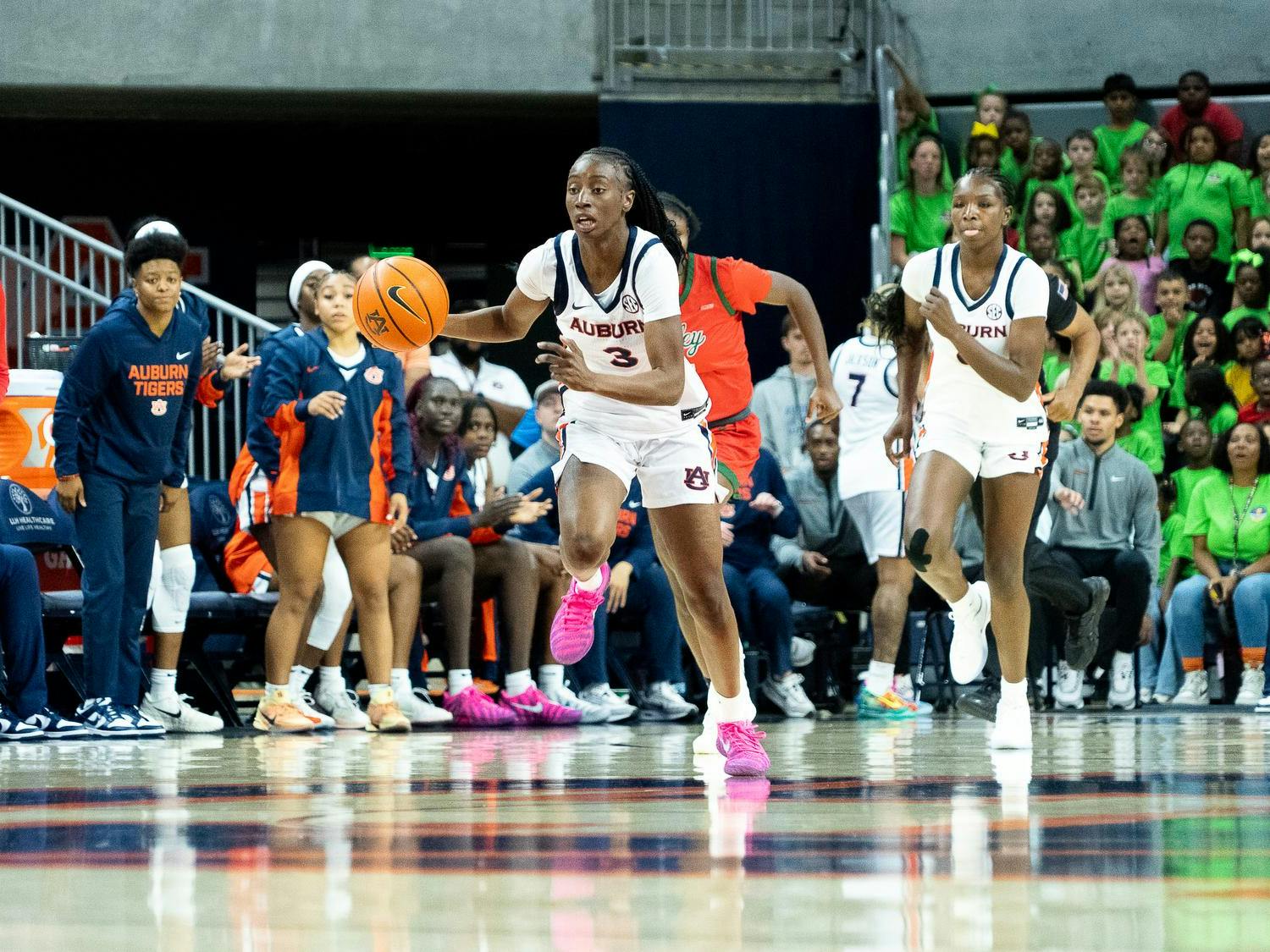 A women’s college basketball player from Auburn, wearing a white uniform, dribbles the ball quickly up the court during a game, with a teammate running alongside her. An opposing player in a red and green uniform trails close behind. The Auburn bench and a crowd of spectators, including a large group wearing bright green shirts, are visible in the background.