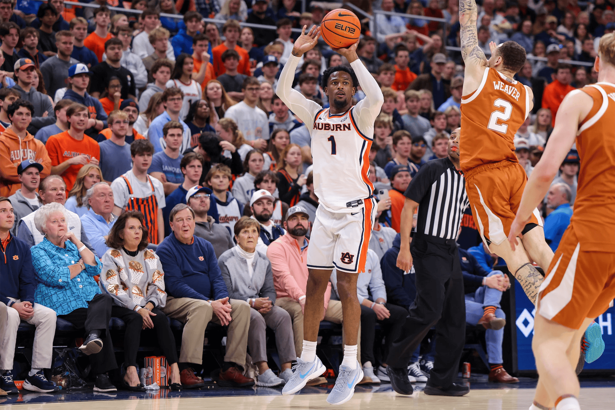 Auburn men’s basketball player in a white No. 1 jersey rises for a jump shot as a Texas defender in a burnt orange uniform contests with an outstretched arm, while a referee and a packed crowd watch from the background.