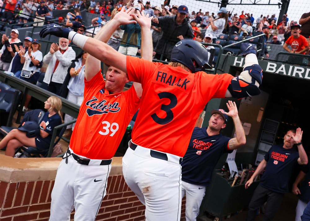 An Auburn baseball player wearing an orange jersey with “FRALICK” and the number 3 on the back leaps into the air near the dugout, celebrating with teammates after a big play. A teammate in an orange jersey raises both hands for a high-five while other players in navy Auburn Tigers gear cheer from the dugout. Fans in the packed stadium stands applaud and reach toward the field, creating an energetic game-day atmosphere.