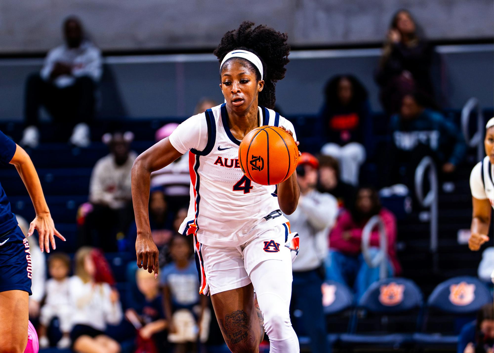 Auburn Women's Basketball guard Kaitlyn Duhon (#4) wears a white headband and a white Auburn Tigers jersey with orange and navy trim as she drives down the court holding a basketball during a game.