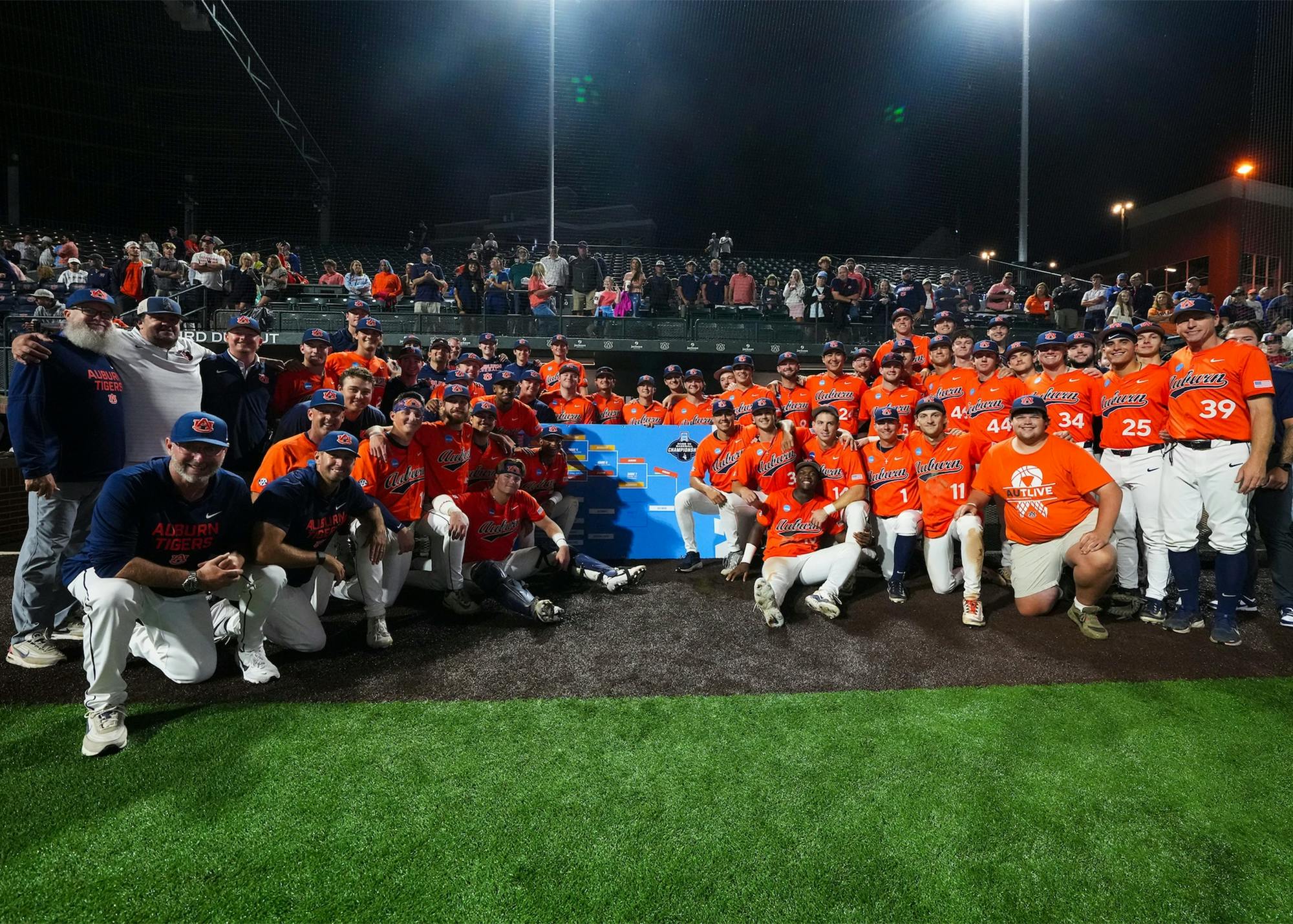 The Auburn baseball team, coaches, and staff pose for a group photo on the field at Plainsman Park following their NCAA Auburn Regional championship victory over Milwaukee. Players in orange jerseys surround the regional bracket while fans stand and cheer in the background under the stadium lights. The win sent Auburn to its second-ever home Super Regional and sixth Super Regional appearance in program history.