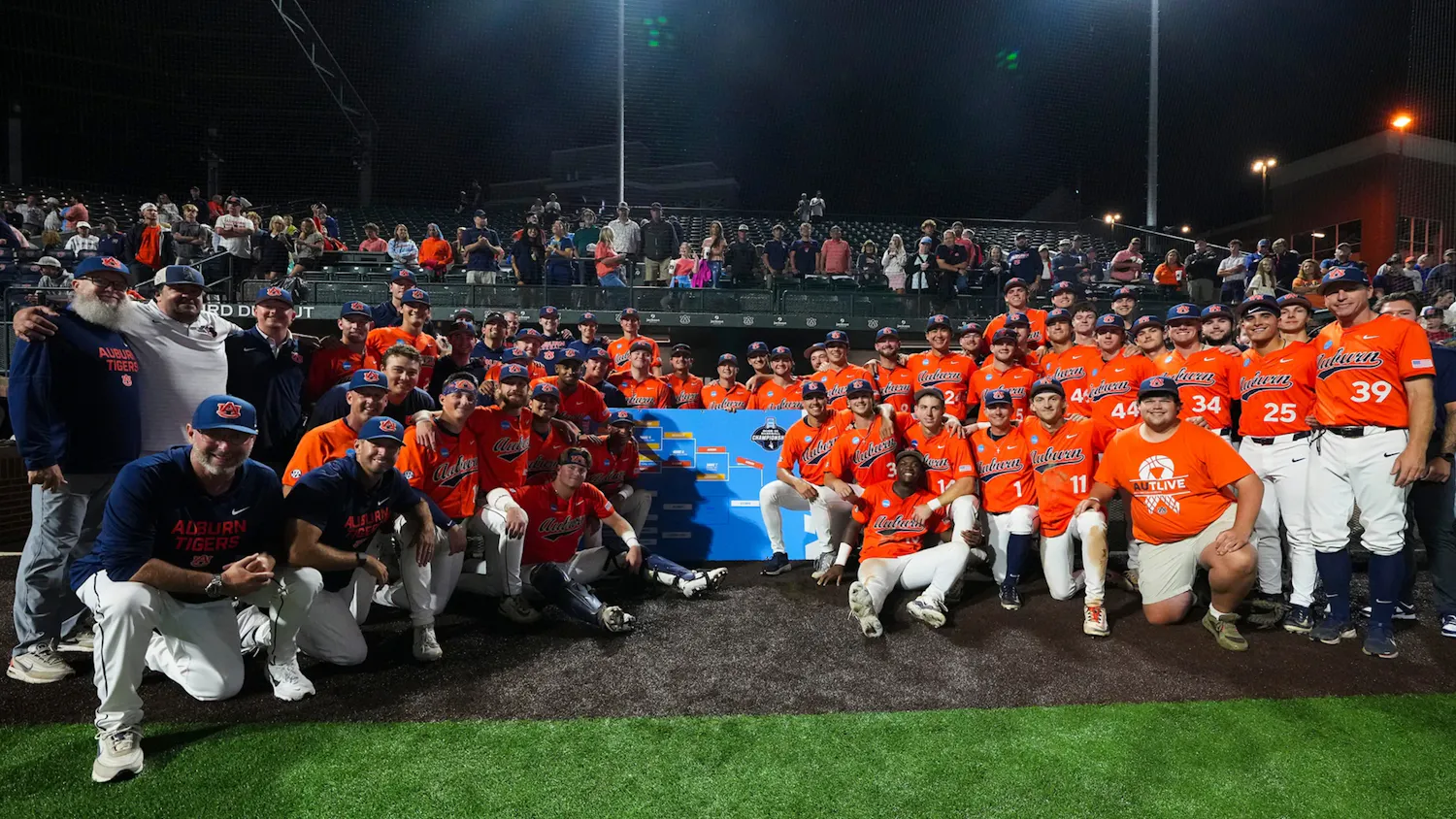 The Auburn baseball team, coaches, and staff pose for a group photo on the field at Plainsman Park following their NCAA Auburn Regional championship victory over Milwaukee. Players in orange jerseys surround the regional bracket while fans stand and cheer in the background under the stadium lights. The win sent Auburn to its second-ever home Super Regional and sixth Super Regional appearance in program history.