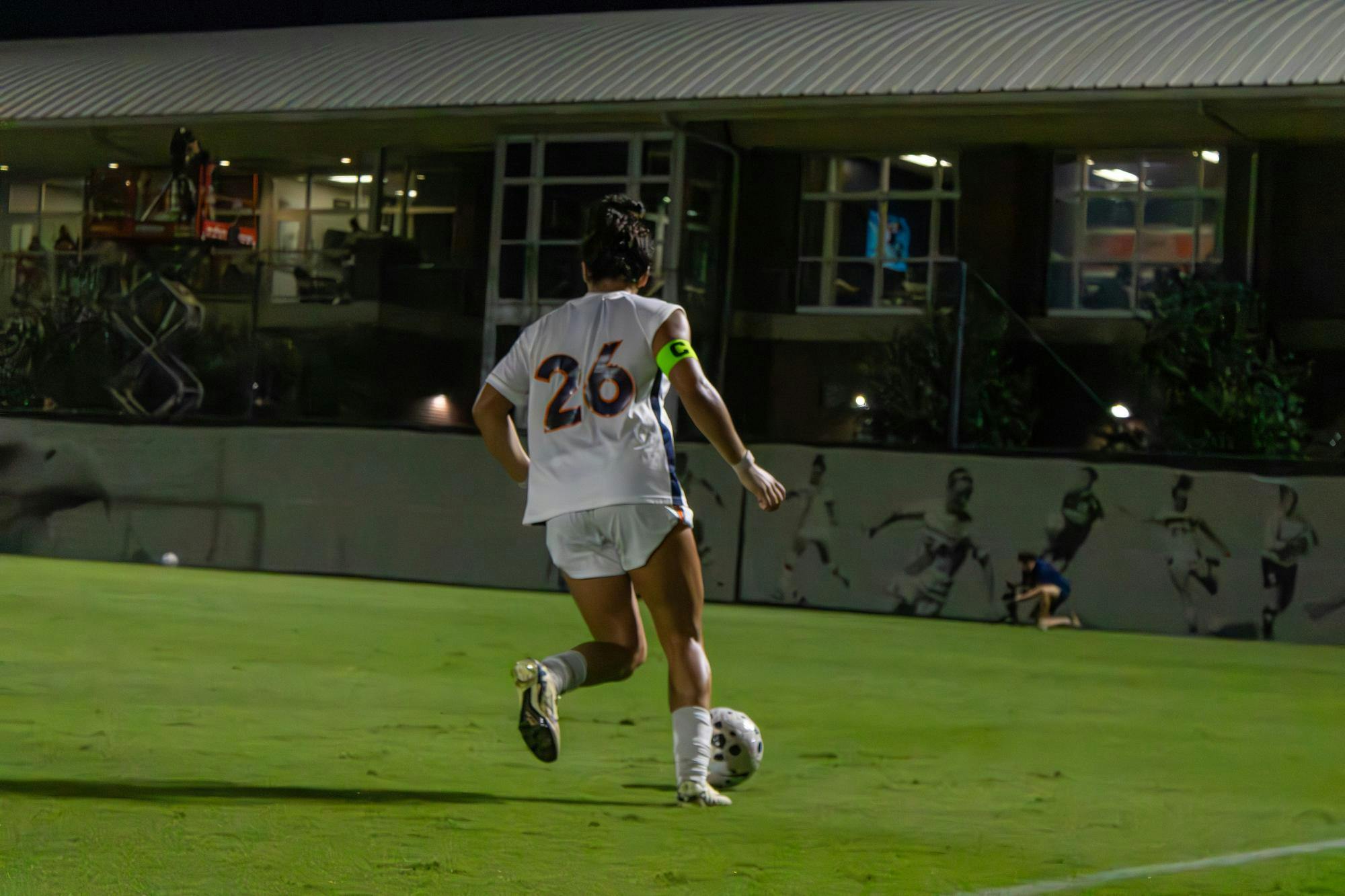 An Auburn soccer player wearing a white uniform dribbles down the right side of a soccer pitch during a night game, illuminated by the floodlights