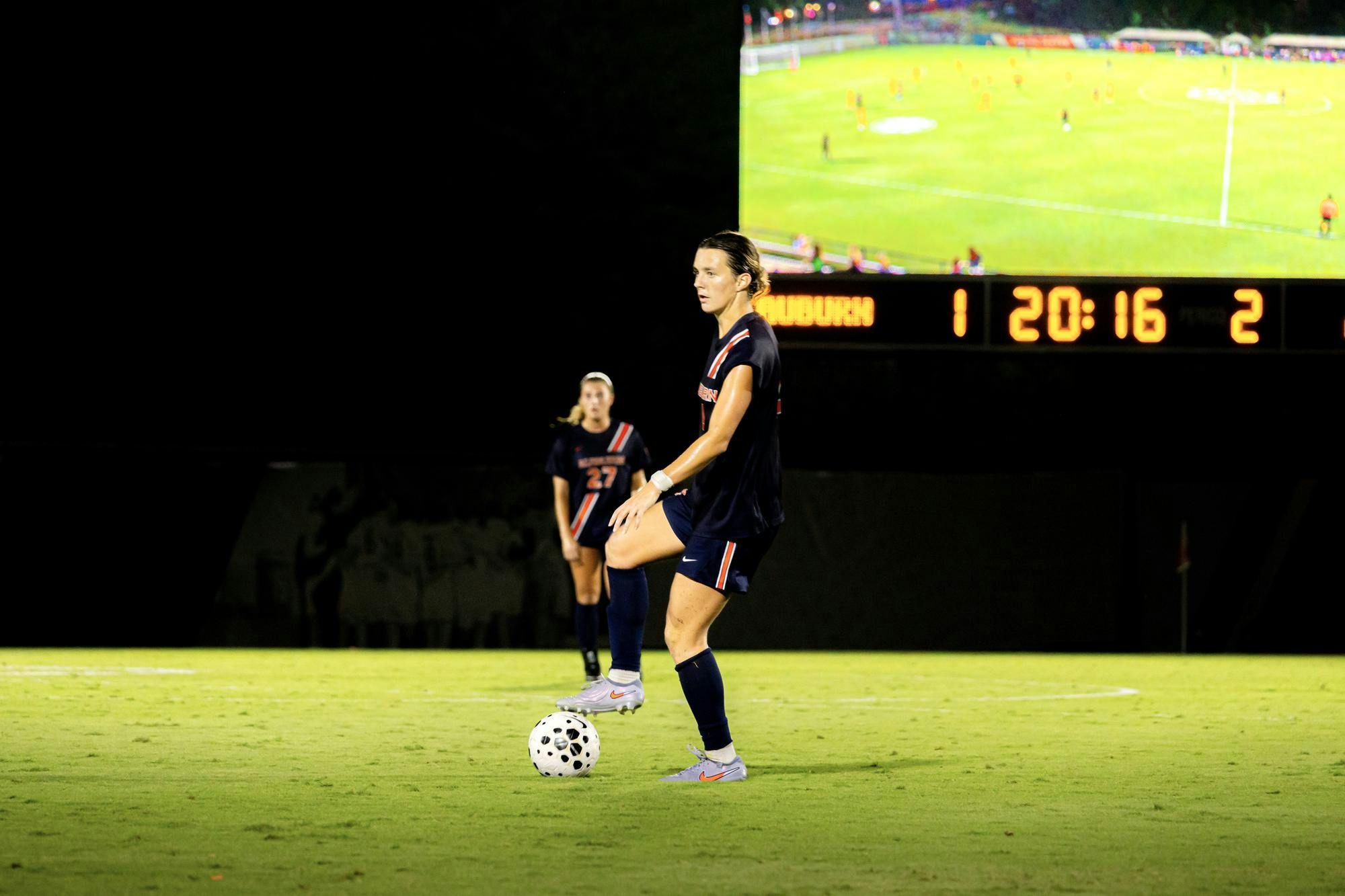 Auburn's Taylor Chism prepares to kick the ball during a night game, with the scoreboard and a live video screen visible in the background. Another teammate stands behind her on the field.