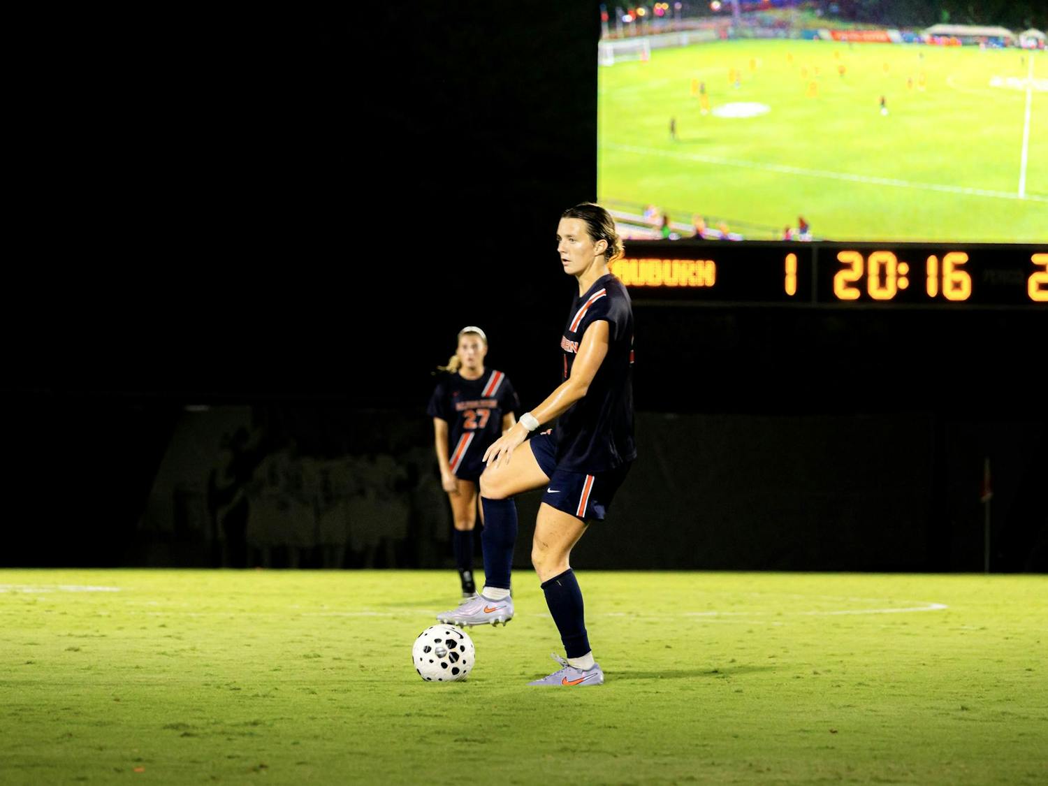 Auburn's Taylor Chism prepares to kick the ball during a night game, with the scoreboard and a live video screen visible in the background. Another teammate stands behind her on the field.