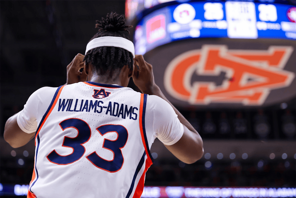 Auburn men’s basketball player wearing a white No. 33 jersey with “Williams-Adams” on the back adjusts his headband while standing on the court, with a large Auburn logo and scoreboard blurred in the background.