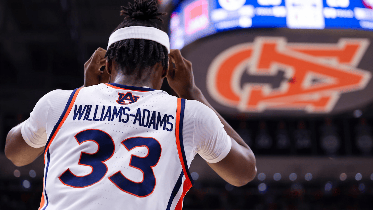 Auburn men’s basketball player wearing a white No. 33 jersey with “Williams-Adams” on the back adjusts his headband while standing on the court, with a large Auburn logo and scoreboard blurred in the background.