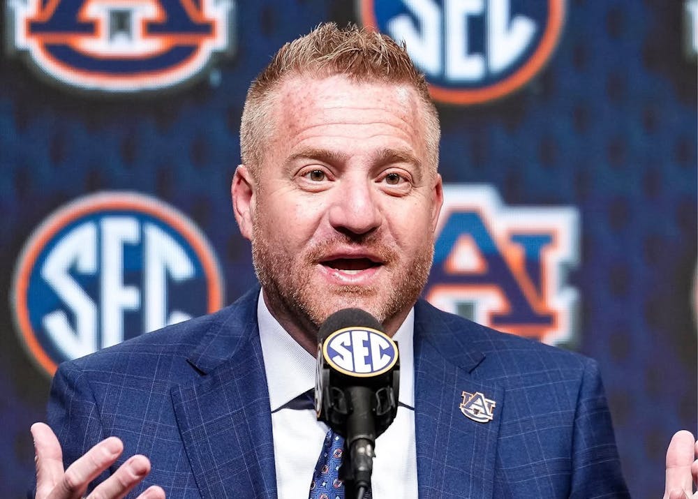 Auburn head coach Alex Golesh speaks at the podium during the 2026 SEC Football Kickoff Media Days, gesturing with both hands while answering questions with SEC and Auburn logos displayed behind him.