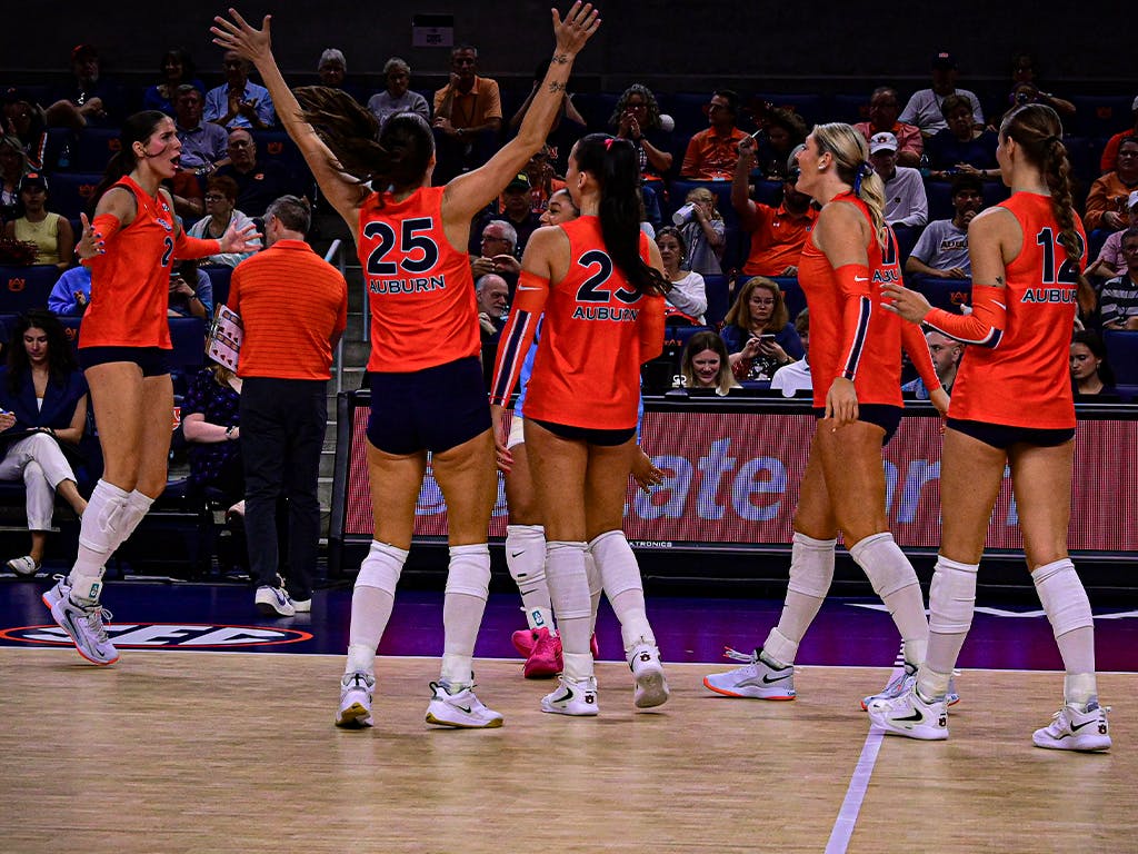 Auburn volleyball players in orange jerseys celebrate a point during a home match as fans cheer in the background.