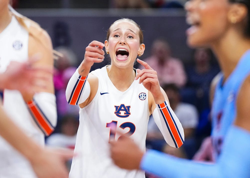 An Auburn volleyball player wearing a white jersey with number 12 gestures and shouts during a match. Her expression appears intense and focused as she communicates with teammates. Other players in Auburn and opposing team uniforms are visible nearby, with a blurred crowd in the background.