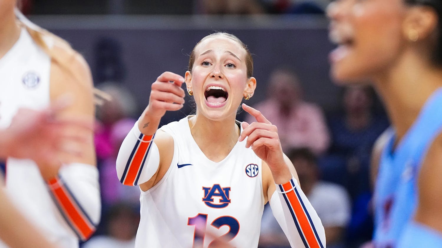 An Auburn volleyball player wearing a white jersey with number 12 gestures and shouts during a match. Her expression appears intense and focused as she communicates with teammates. Other players in Auburn and opposing team uniforms are visible nearby, with a blurred crowd in the background.