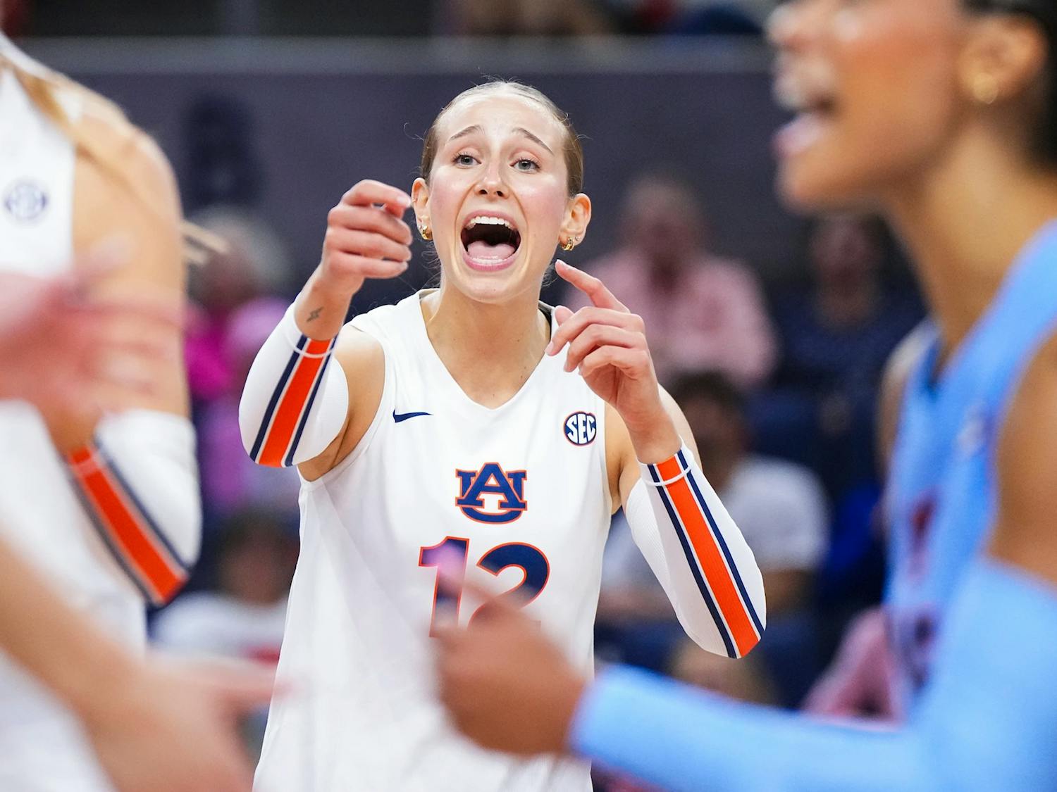 An Auburn volleyball player wearing a white jersey with number 12 gestures and shouts during a match. Her expression appears intense and focused as she communicates with teammates. Other players in Auburn and opposing team uniforms are visible nearby, with a blurred crowd in the background.