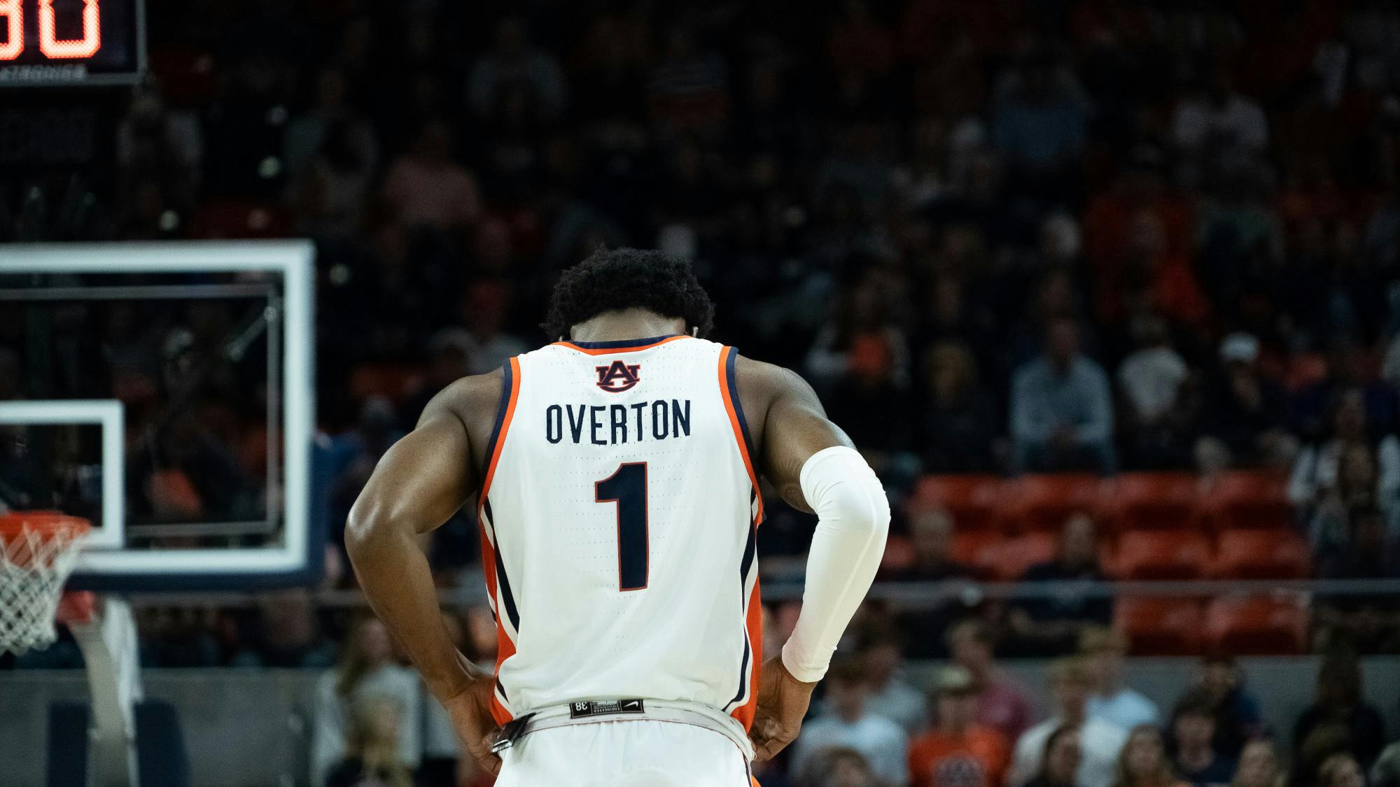 Back view of an Auburn men’s basketball player standing on the court with hands on hips, wearing a white jersey with “Overton” and the number 1, as the crowd and arena seats blur in the background.

