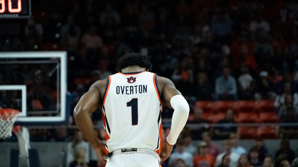 Back view of an Auburn men’s basketball player standing on the court with hands on hips, wearing a white jersey with “Overton” and the number 1, as the crowd and arena seats blur in the background.
