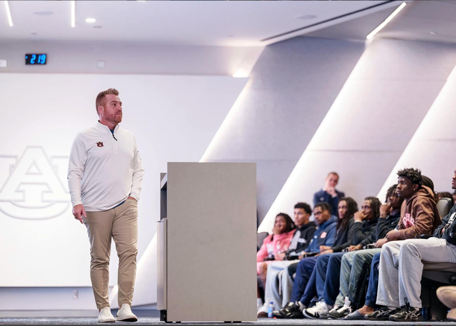 Auburn head football coach Alex Golesh stands in front of his team during a meeting, speaking to players seated in an auditorium. The Auburn logo is visible on the wall behind him.