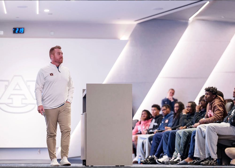 Auburn head football coach Alex Golesh stands in front of his team during a meeting, speaking to players seated in an auditorium. The Auburn logo is visible on the wall behind him.