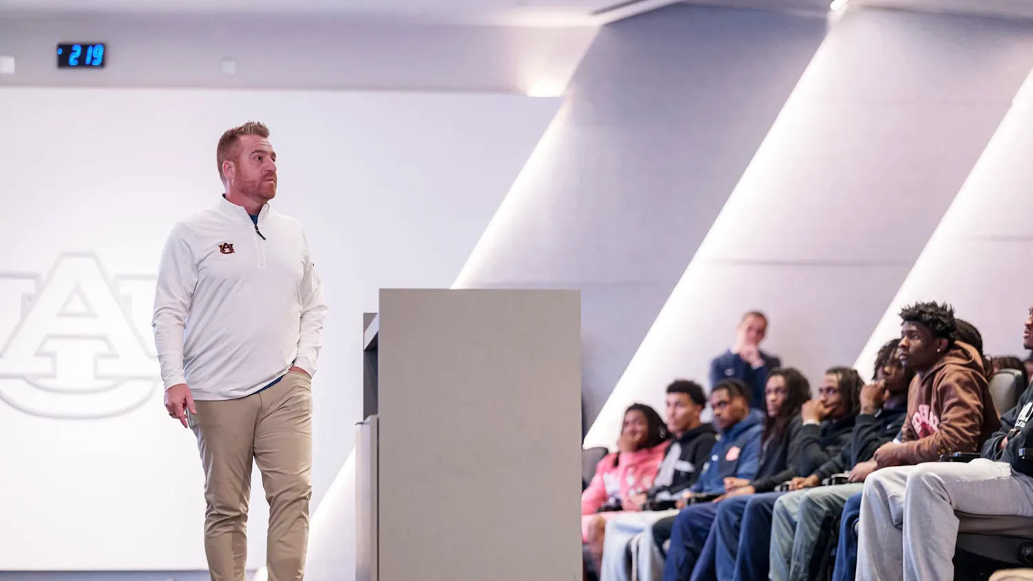 Auburn head football coach Alex Golesh stands in front of his team during a meeting, speaking to players seated in an auditorium. The Auburn logo is visible on the wall behind him.