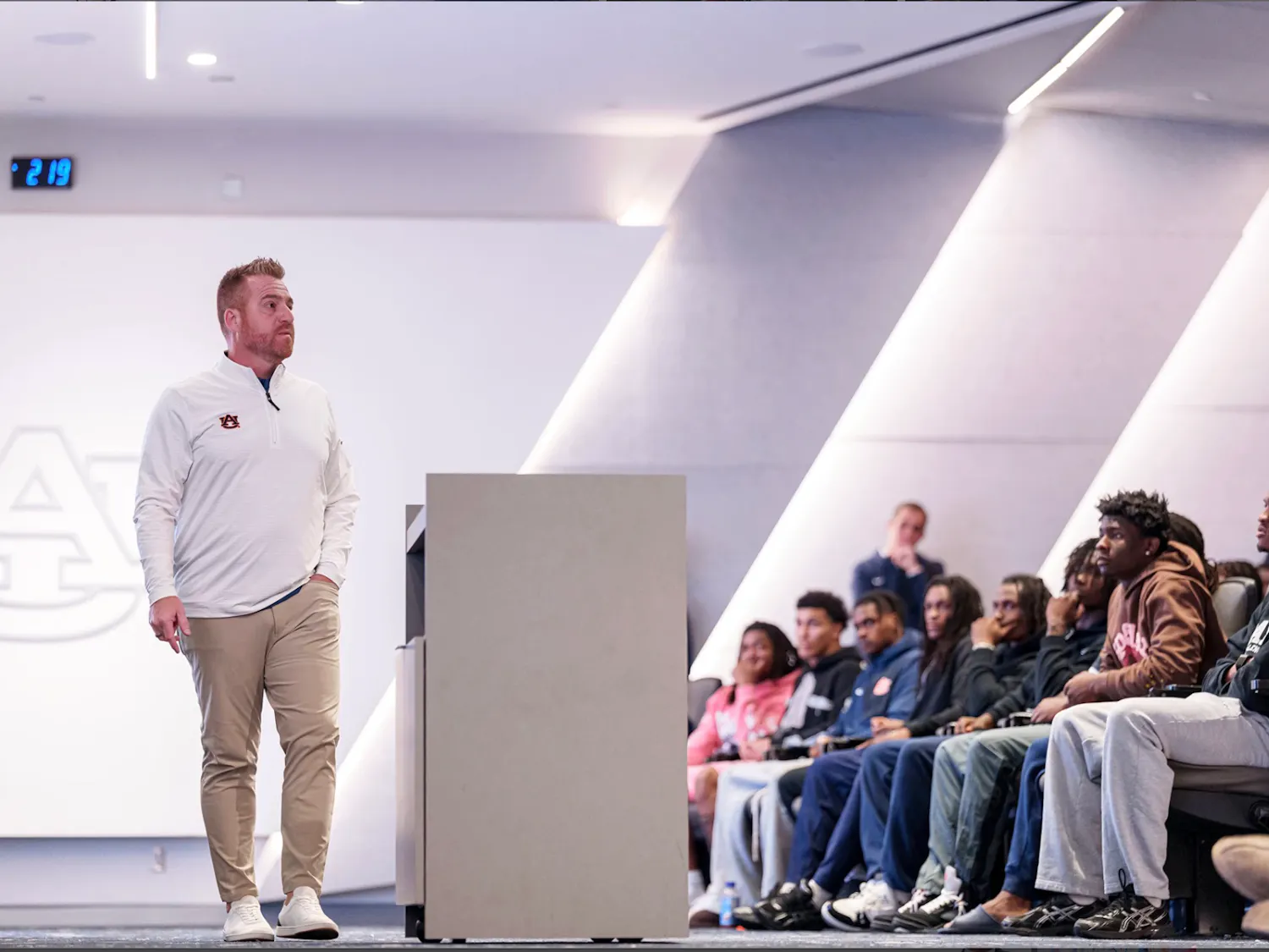Auburn head football coach Alex Golesh stands in front of his team during a meeting, speaking to players seated in an auditorium. The Auburn logo is visible on the wall behind him.
